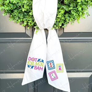 Könnte beinhalten: Ein weißes Stoffband mit dem farbigen Text DOT CRACK BAM und drei Mahjong-Stein-Illustrationen in Pink, Blau und Grün. Das Band ist zu einem Knoten gebunden und hängt mit zwei langen Enden herab, die detaillierte Grafiken zum Spiel zeigen.