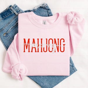 May include: A light pink sweatshirt with the word "MAHJONG" in red block letters. The word "LOVE" is written in white script over the "O" in "MAHJONG". The sweatshirt is folded over a pair of blue jeans.