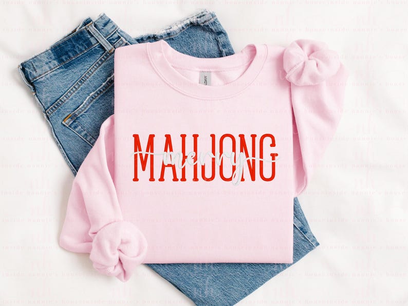 May include: A light pink sweatshirt with the word "MAHJONG" in red block letters. The word "LOVE" is written in white script over the "O" in "MAHJONG". The sweatshirt is folded over a pair of blue jeans.