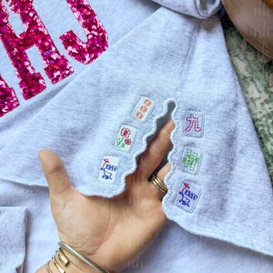 Op de afbeelding: Lichtgrijs sweatshirt met roze pailletten letters "HAJ". De onderkant heeft een geschulpte rand met geborduurde patches van Mahjong-tegels en Chinese karakters.