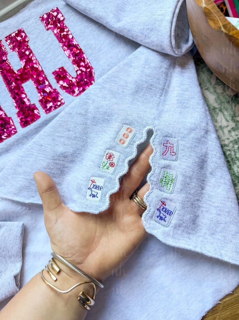 Op de afbeelding: Lichtgrijs sweatshirt met roze pailletten letters "HAJ". De onderkant heeft een geschulpte rand met geborduurde patches van Mahjong-tegels en Chinese karakters.