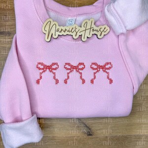 Puede incluir: Sudadera rosa claro con tres lazos rojos bordados. Los puños son blancos. Una placa de madera con la inscripción "Nannie's House" está adjunta.