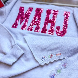 Op de afbeelding: Een lichtgrijs sweatshirt met het woord "MAHJ" in grote, glinsterende roze letters op de voorkant. De zoom van de trui heeft geborduurde patches met kleurrijke ontwerpen. Het sweatshirt is een casual kledingstuk.