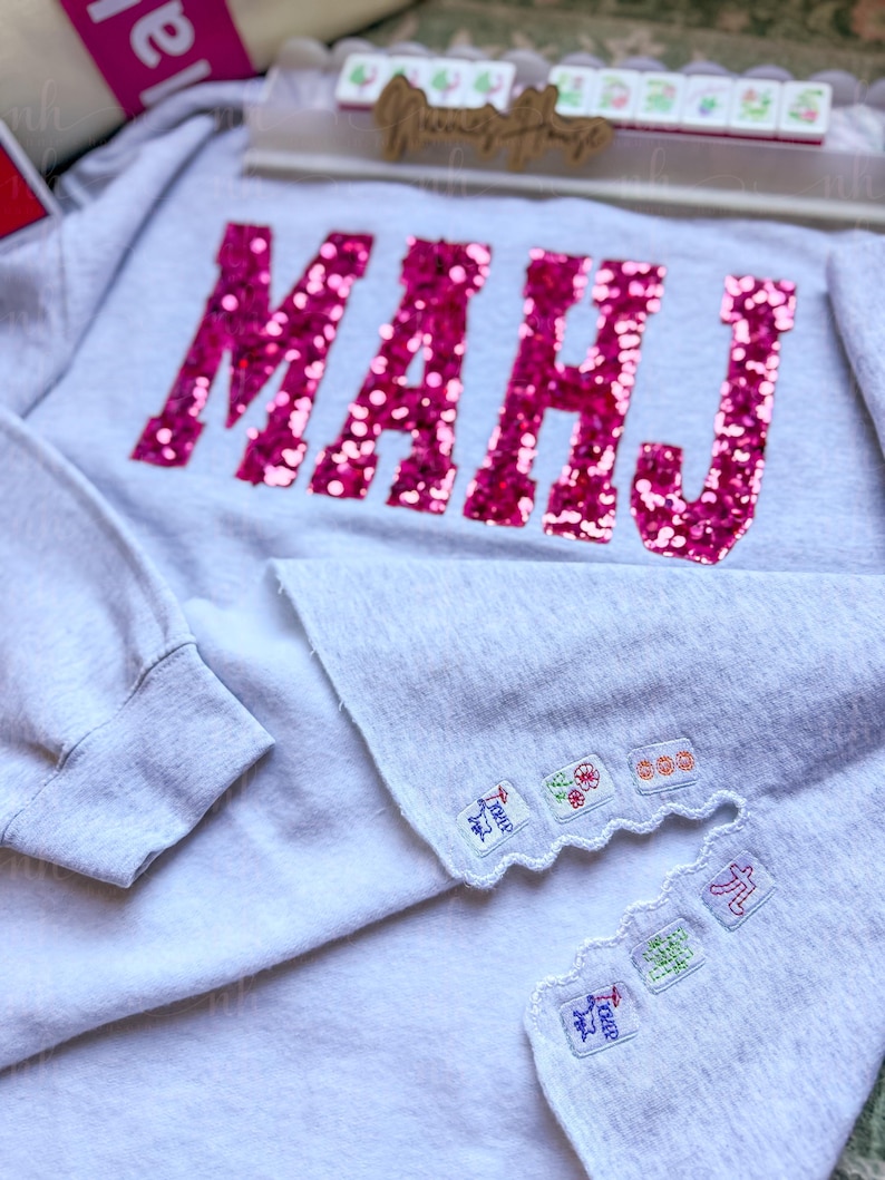 Op de afbeelding: Een lichtgrijs sweatshirt met het woord "MAHJ" in grote, glinsterende roze letters op de voorkant. De zoom van de trui heeft geborduurde patches met kleurrijke ontwerpen. Het sweatshirt is een casual kledingstuk.