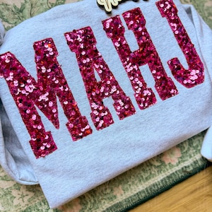 Op de afbeelding: Een lichtgrijze sweatshirt met de letters "MAHJ" in grote, sprankelende roze pailletten. De sweatshirt is opgevouwen op een groen en wit oppervlak met een patroon, met een houten bordje met de tekst "Nannie's House".