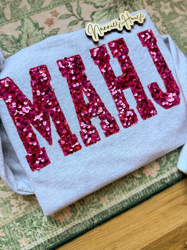 Op de afbeelding: Een lichtgrijze sweatshirt met de letters "MAHJ" in grote, sprankelende roze pailletten. De sweatshirt is opgevouwen op een groen en wit oppervlak met een patroon, met een houten bordje met de tekst "Nannie's House".