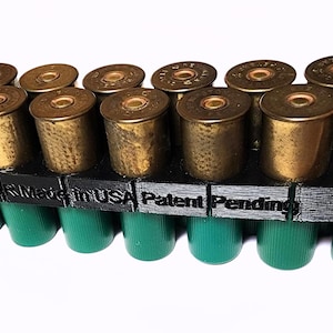 Puede incluir: Un portacartuchos de escopeta de plástico negro con 12 cartuchos de escopeta. El portacartuchos tiene el texto "MIL 12 GA 10PK Made in USA Patent Pending" impreso en él.