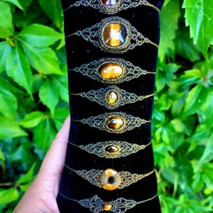 Könnte beinhalten: Eine Auswahl an Makramee-Armbändern mit verschiedenen Edelsteinen, darunter Tigerauge, präsentiert auf einem schwarzen Samtkissen. Im Hintergrund sind grüne Blätter zu sehen, die einen farblichen Kontrast bilden.