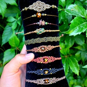 Könnte beinhalten: Eine Sammlung handgefertigter Makramee-Armbänder in verschiedenen Farben, darunter Braun, Grün und Rot. Einige Armbänder haben Perlen und Edelsteine. Die Armbänder werden auf einem schwarzen Samtuntergrund präsentiert.