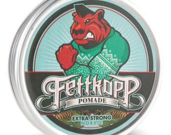 May include: A silver tin with a blue and green label featuring a cartoon bear wearing a green sweater with a white snowflake pattern. The label reads "Fettkopp Pomade Extra Strong Nordic".