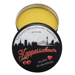 Könnte beinhalten: Eine schwarze Dose mit gelbem Deckel. Die Dose hat ein weiß-schwarzes Stadtbild-Design mit dem Text "Feinstes Düsseldorfer Haarfett - de zwedde -" und "Kappesschmöör" in Rot. Die Dose hat auch ein rotes Herz und den Text "Aus dem des Rheinlands!" in Weiß.