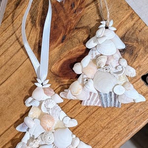 May include: Two white seashell Christmas tree ornaments. The ornaments are made of various sizes and shapes of seashells, with a white ribbon and twine for hanging.