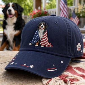 May include: A navy blue baseball cap featuring an embroidered Bernese Mountain Dog wrapped in an American flag on the front. The brim displays small white paw prints, stars, and red and white stripes. A paw print with a red star is on the side.
