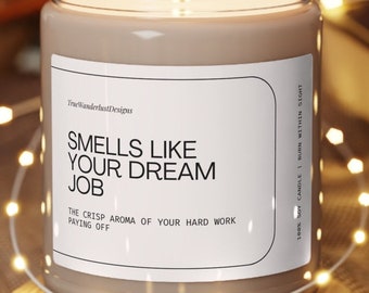 Op de afbeelding: Een bruine sojawas-kaars met een wit label waarop staat "Smells Like Your Dream Job. The crisp aroma of your hard work paying off." De kaars staat op een houten onderzetter.