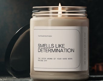 Op de afbeelding: Een beige sojawas kaars met een wit label waarop staat "Smells Like Determination" en "The Crisp Aroma of Your Hard Work Paying Off". De kaars zit in een glazen pot met een zwarte deksel. Het label bevat ook de tekst "TrueWanderlustDesigns" en "100% Soy Candle | Burn Within Sight".