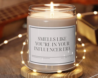 Op de afbeelding: Een beige kaars met een wit label waarop staat "Smells like you're in your influencer era". De kaars is aangestoken en omringd door lichtsnoeren.