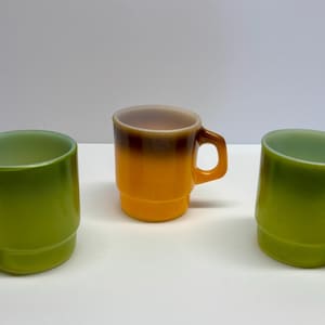 Puede incluir: Tres tazas de café vintage. Dos son verdes con interior verde claro, y una es naranja con la parte superior marrón. Las tazas tienen asa y diseño apilado. Son de vidrio.