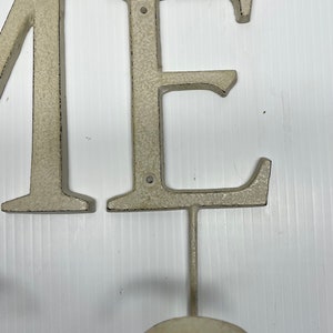 Puede incluir: Letras de metal blanco que deletrean "ME" con un gancho unido a la letra "E".