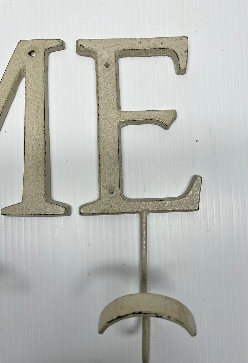 Puede incluir: Letras de metal blanco que deletrean "ME" con un gancho unido a la letra "E".