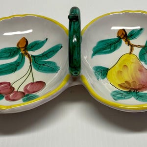 Puede incluir: Un plato de servir de cer&aacute;mica blanca con dos secciones, cada una con un dise&ntilde;o de fruta pintado a mano. Un lado tiene una pera y el otro tiene cerezas. El plato tiene un borde amarillo y un asa verde.