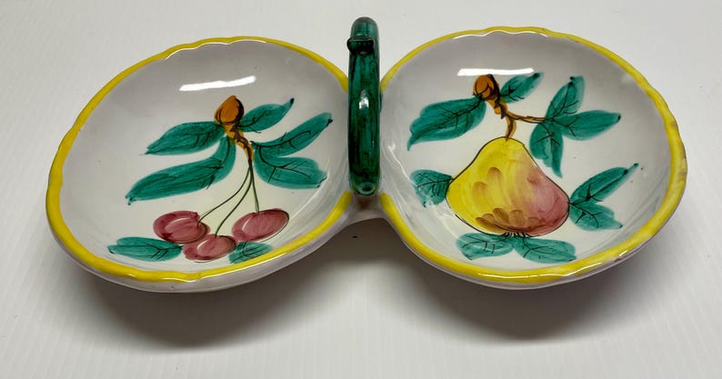 Puede incluir: Un plato de servir de cer&aacute;mica blanca con dos secciones, cada una con un dise&ntilde;o de fruta pintado a mano. Un lado tiene una pera y el otro tiene cerezas. El plato tiene un borde amarillo y un asa verde.