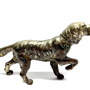 May include: A detailed, antique-style dog figurine in a walking pose. The sculpture is metallic with a mix of silver and bronze tones, showcasing intricate fur and muscle details. The dog is depicted with one paw raised, suggesting movement.