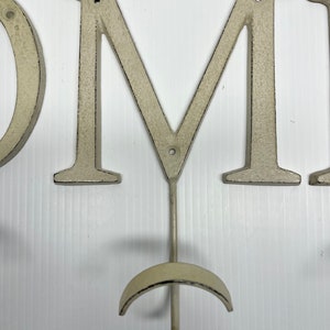 Puede incluir: Letras de metal blanco que deletrean "HOME" con un gancho en la "M". Las letras est&aacute;n desgastadas y tienen un aspecto r&uacute;stico.