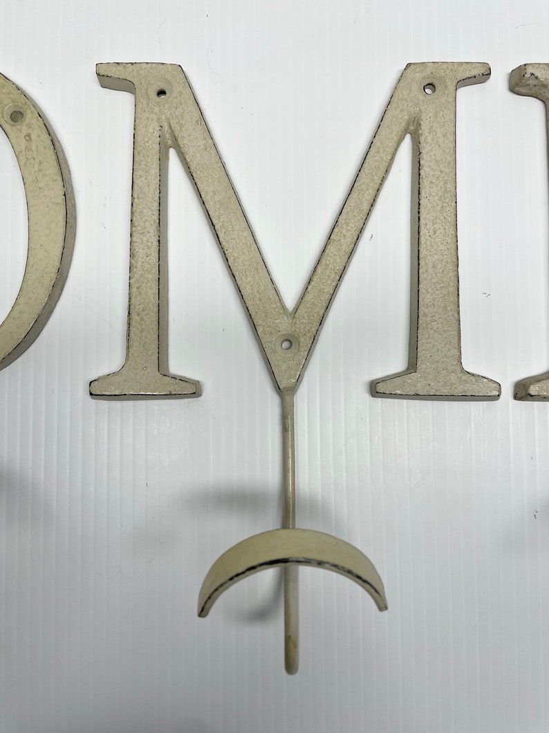 Puede incluir: Letras de metal blanco que deletrean "HOME" con un gancho en la "M". Las letras est&aacute;n desgastadas y tienen un aspecto r&uacute;stico.