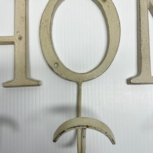 Puede incluir: Un letrero de metal blanco "HOME" con un gancho en la "O". El letrero est&aacute; desgastado y tiene un aspecto r&uacute;stico.