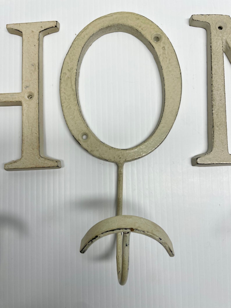 Puede incluir: Un letrero de metal blanco "HOME" con un gancho en la "O". El letrero est&aacute; desgastado y tiene un aspecto r&uacute;stico.