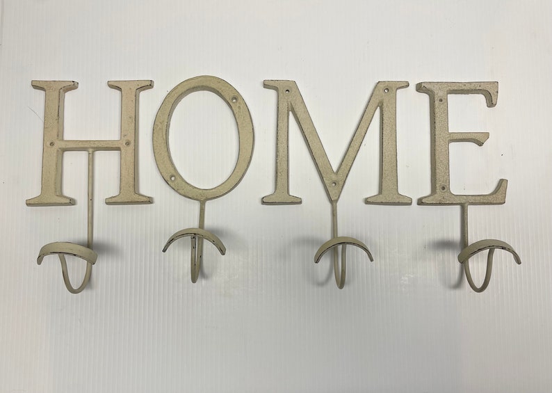 Puede incluir: Letrero HOME de metal blanco con ganchos para colgar objetos. Cada letra tiene un gancho debajo.