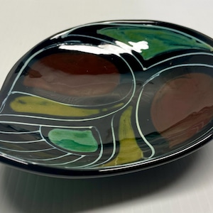 Puede incluir: Un plato de cerámica negro en forma de hoja con acabado brillante. El plato presenta un diseño de formas marrones, verdes y amarillas delineadas en blanco. El plato mide aproximadamente 25 cm de largo y 15 cm de ancho, perfecto para servir aperitivos o como pieza decorativa.