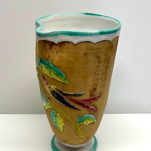 May include: A decorative vase with a textured, golden-brown exterior. It features a floral design with green, yellow, blue, and red accents. The rim is white with a scalloped edge and a teal border. The base is also teal.