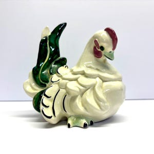 May include: Cream-colored ceramic hen figurine with a green and black tail. The hen has a red comb and wattle, and black accents on its feet. The figurine is a decorative item, likely for display.