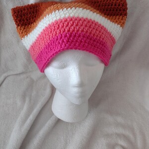 May include: A handmade, crocheted beanie in a striped pattern of pink, white, orange, and brown. The hat is displayed on a white foam head. The beanie's design features a series of horizontal stripes, showcasing a gradient of colors.