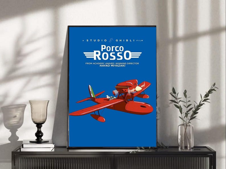 Op de afbeelding: Een ingelijste poster van de animatiefilm "Porco Rosso" van Studio Ghibli. De poster toont een rood watervliegtuig tegen een blauwe achtergrond, met de titel van de film in het wit. De tekst "FROM ACADEMY AWARD-WINNING DIRECTOR HAYAO MIYAZAKI" is ook opgenomen.