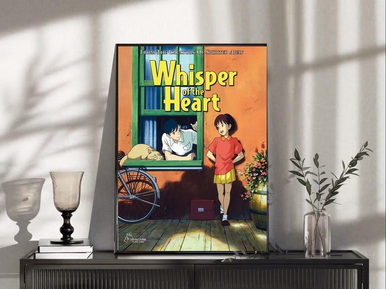 May include: A framed poster featuring the anime film "Whisper of the Heart." The artwork depicts two characters, a boy and a girl, with a cat, a bicycle, and the film's title in yellow. The background is a warm orange and green.