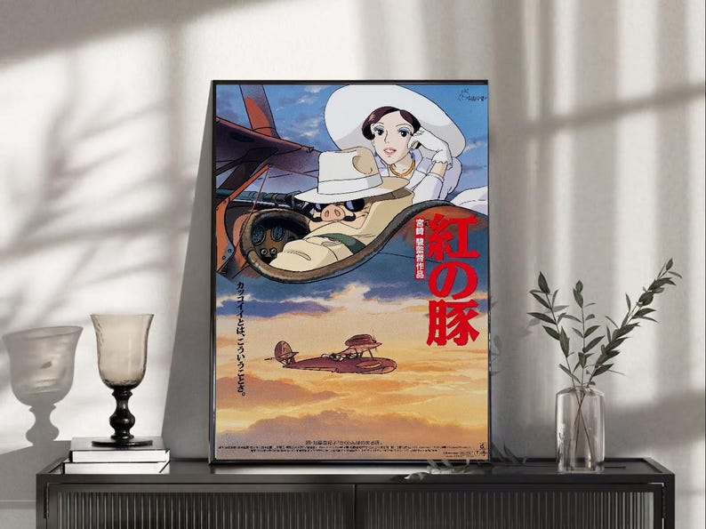 May include: Framed poster featuring a vibrant illustration from the animated film "Porco Rosso." The artwork depicts a pig-like character in a biplane, with a woman in a white hat. Japanese text is visible. The background shows a sunset sky.