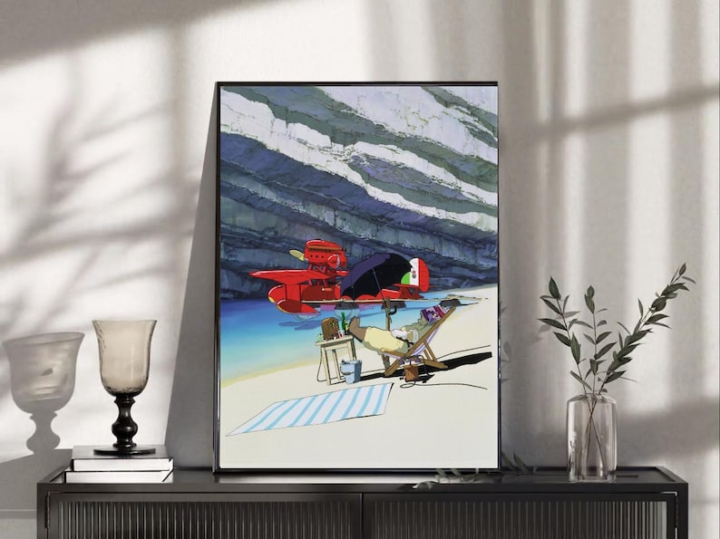 May include: Framed art print featuring a vibrant illustration of a red seaplane on the water near a beach. A person relaxes in a beach chair, with a striped towel and small table nearby. The background shows a rocky cliff and blue water.