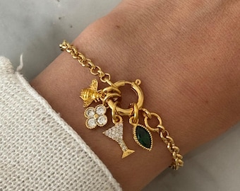 Op de afbeelding: Een goudkleurige schakelarmband met een ronde sluiting waaraan vier verschillende bedels hangen: een kleine bij, een klaverblad met doorzichtige steentjes, een silhouet van een martiniglas bezet met steentjes en een donkergroene, markiesgeslepen steen.