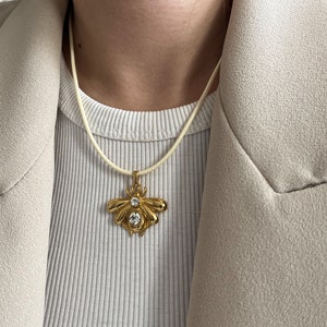 Könnte beinhalten: Eine goldene Bienen-Anhänger-Kette mit klaren Kristall-Akzenten hängt an einer cremefarbenen Schnur.