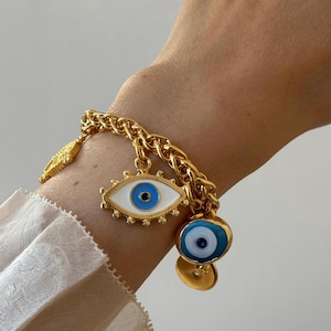 May include: Gold-toned charm bracelet featuring a chain link design. The bracelet has several charms, including an eye-shaped charm with blue and white enamel, a small gold leaf, and a round charm with a blue eye design.