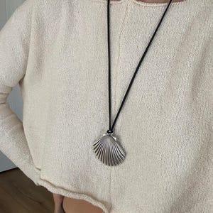 Peut inclure: Un pendentif en forme de coquillage argenté est suspendu à un cordon noir fin. Le pendentif présente un design détaillé en éventail avec des côtes. Le collier est porté sur un haut en maille texturée crème à manches longues et ourlet brut.