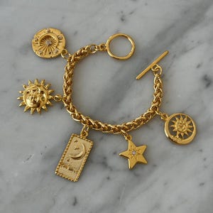 Puede incluir: Pulsera dorada con colgantes de temática celestial. La pulsera incluye colgantes con forma de sol, luna y estrellas, con un cierre de palanca. Los colgantes presentan diseños detallados y pequeños cristales. La pulsera está sobre una superficie de mármol.