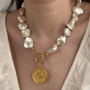 May include: A gold necklace with a large, ornate coin pendant and a chunky chain. The chain is made of irregular, white baroque pearls.