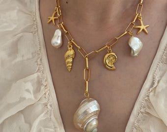 May include: Gold-toned necklace featuring a chain design with various seashell and starfish charms. The necklace includes a large, pearlescent seashell pendant and several smaller shell and starfish accents. The necklace is worn against a cream-colored top.