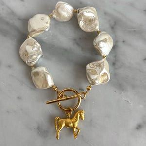 May include: A white pearl bracelet with a gold horse charm and a toggle clasp.