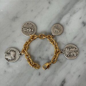 May include: Gold-tone chain bracelet with four silver-colored coin charms. Each coin features a different embossed design. The bracelet has a lobster clasp closure. The bracelet is displayed on a marble surface.