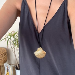 May include: A gold-colored seashell pendant necklace with a black cord. The pendant is a detailed replica of a scallop shell. The necklace is worn over a dark gray V-neck dress.