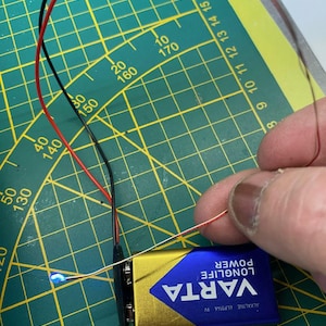 Könnte beinhalten: Nahaufnahme einer goldenen VARTA LONGLIFE 9V Batterie mit einem roten und schwarzen Kabel, das daran angeschlossen ist. Die Batterie befindet sich auf einer grünen Schneidematte mit weißen Markierungen.